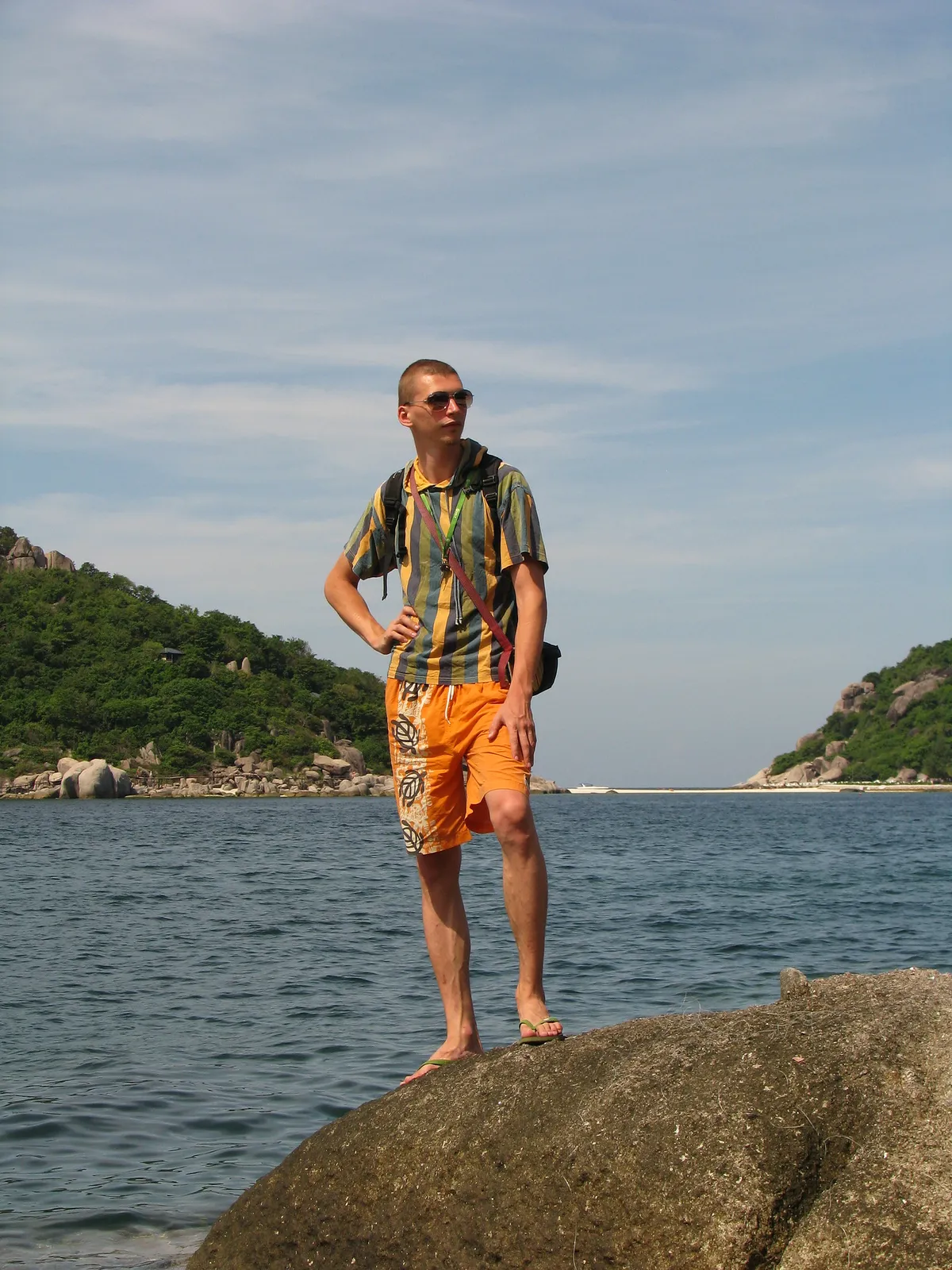 A man in sunglasses, a striped shirt and orange shorts stands barefoot on a large rock above the sea, green islands behind him