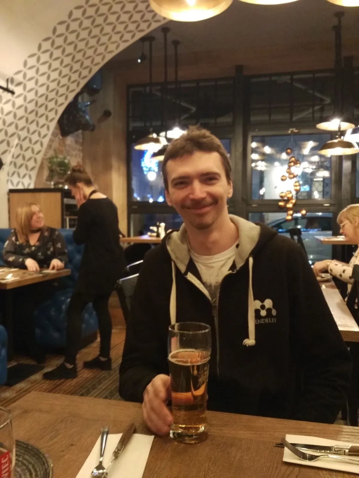 A smiling man in a dark hoodie sits at a wooden table in a restaurant with a glass of beer, arched ceiling and warm lamps behind him