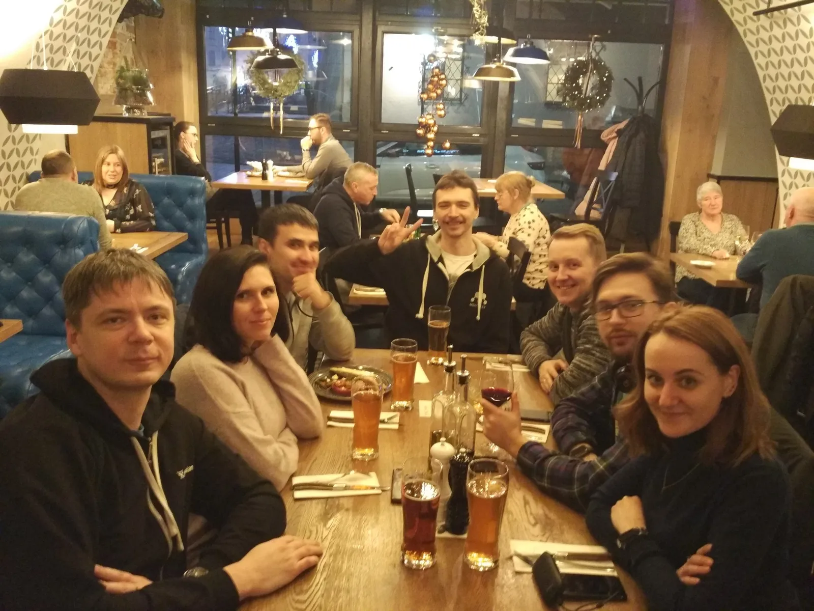 Seven people around a wooden restaurant table with glasses of beer, smiling at the camera, one of them raising a glass, a crowded dining room with festive lights behind