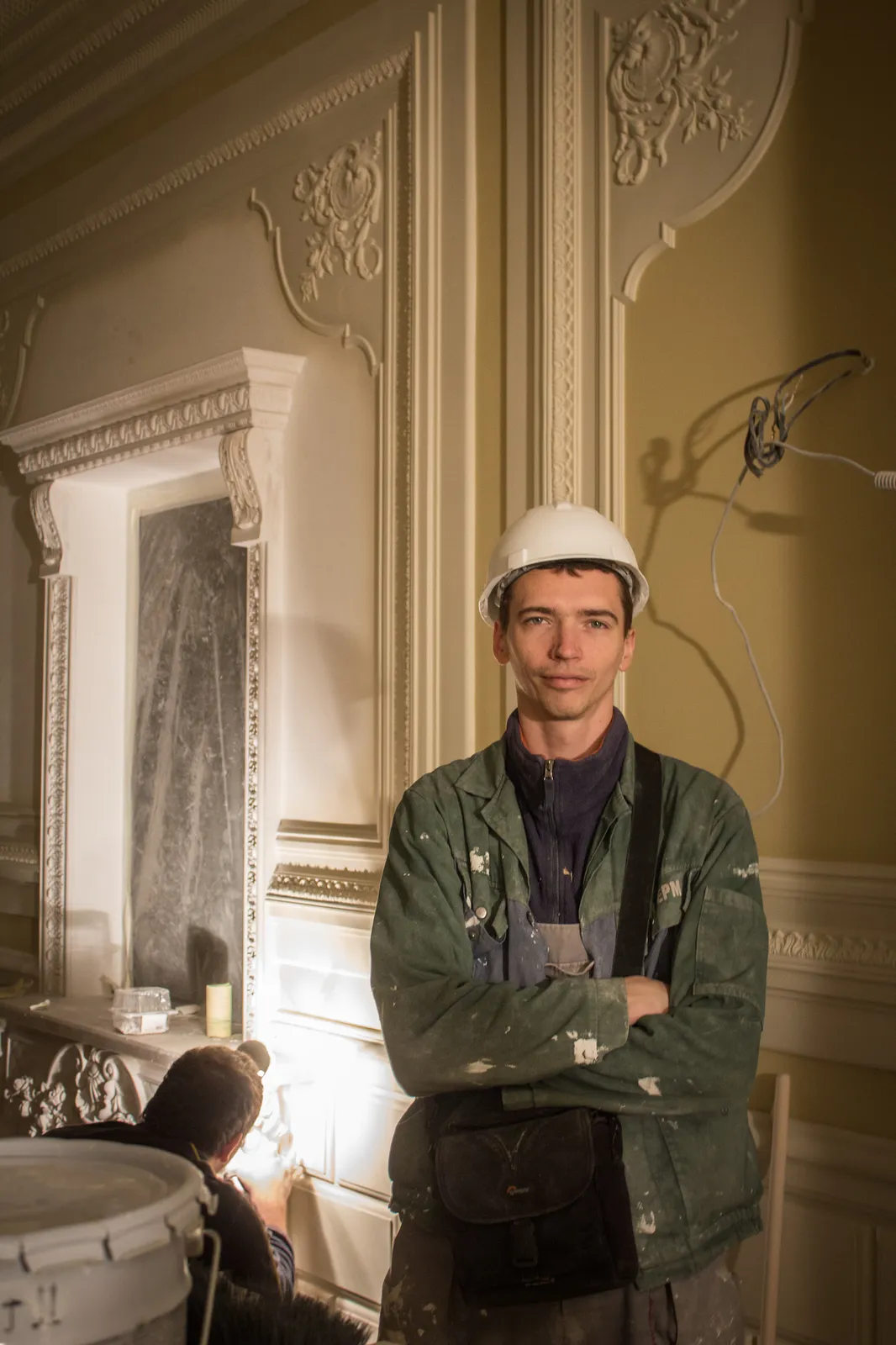 A man in a white hard hat and a green work jacket stands with arms folded in a room with ornate cream stucco walls and a carved door frame