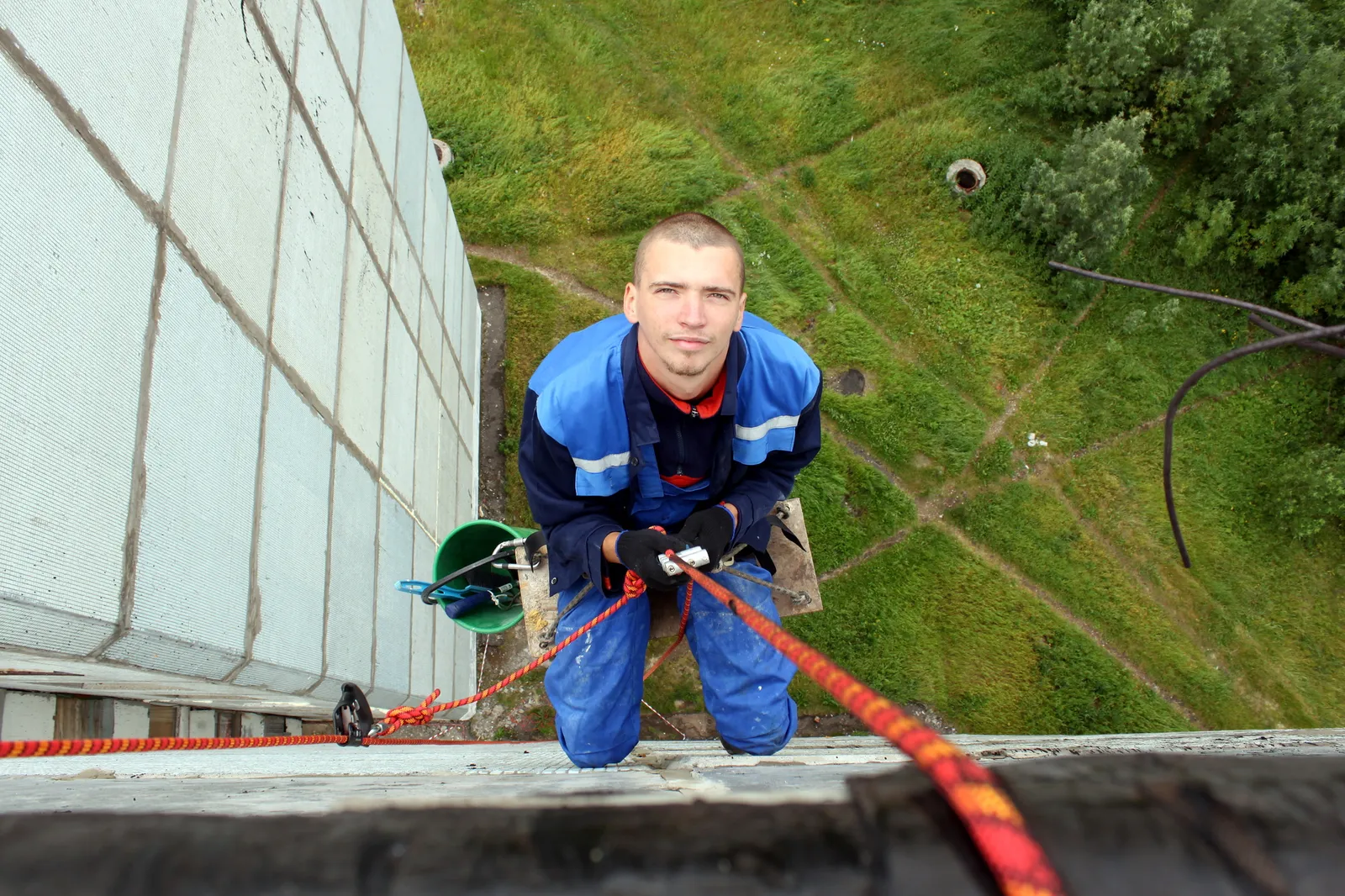 Looking straight down a white panel facade: a man in a blue work suit and gloves hangs on red ropes, smiling up at the camera, a bucket at his side and green grass far below