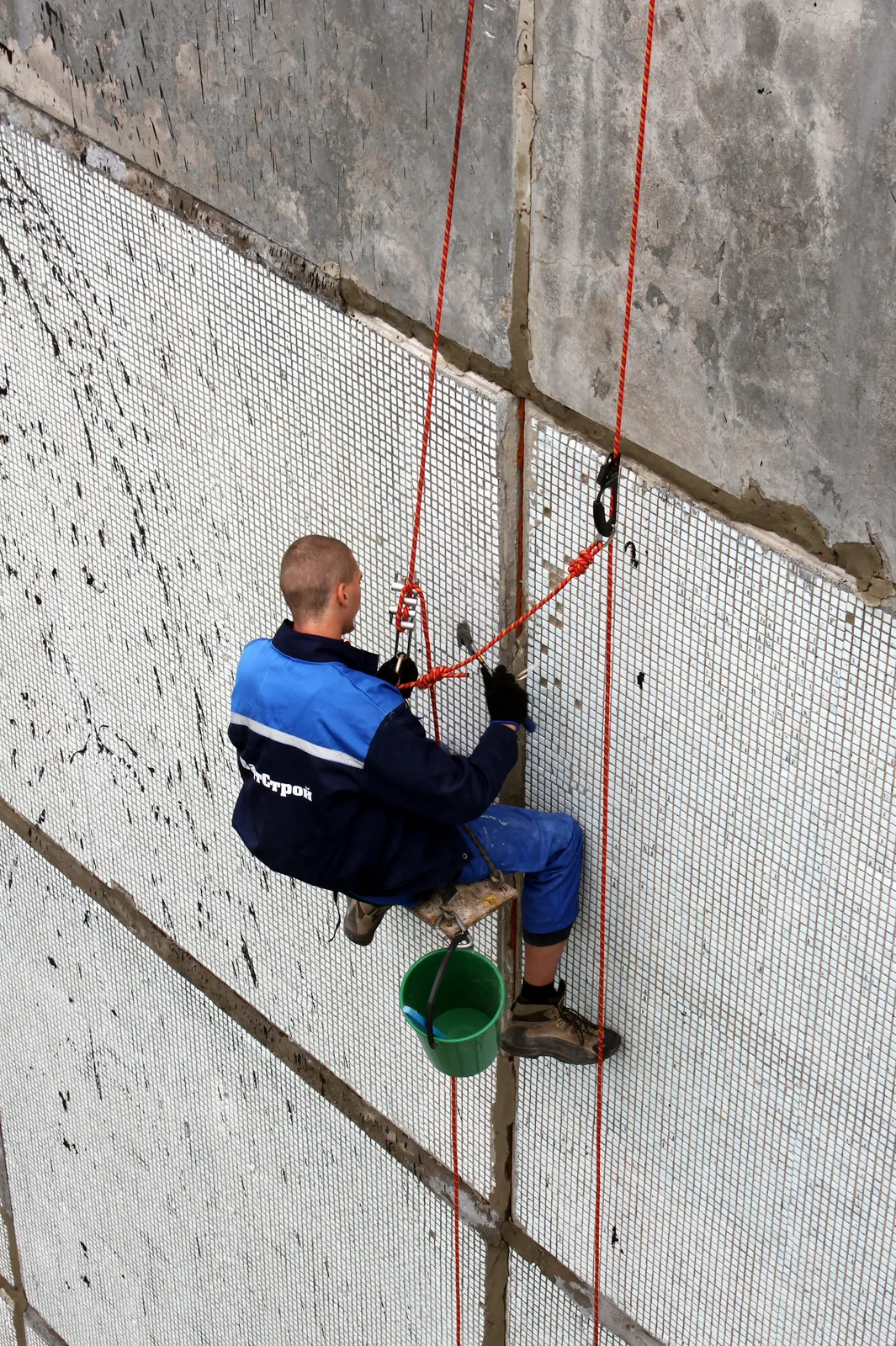 A climber in a blue jacket sits on a board hanging from red ropes against a concrete panel wall covered in white mesh, a green bucket beside him